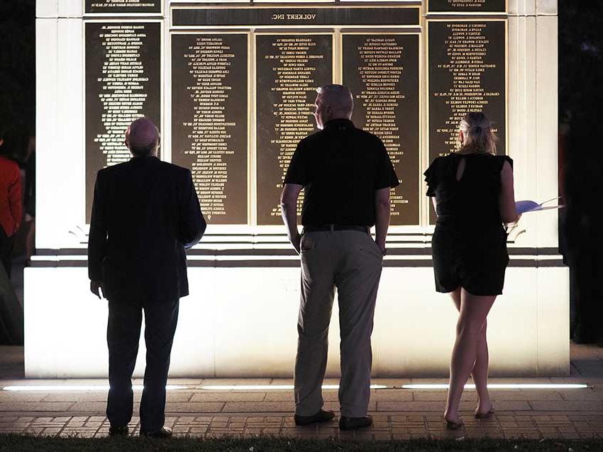 Three people in front of wall of honor.